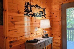 Interior de uma cabana de madeira com uma luminária na parede, uma pequena mesa com uma estatueta de urso e uma porta que dá para uma varanda externa.