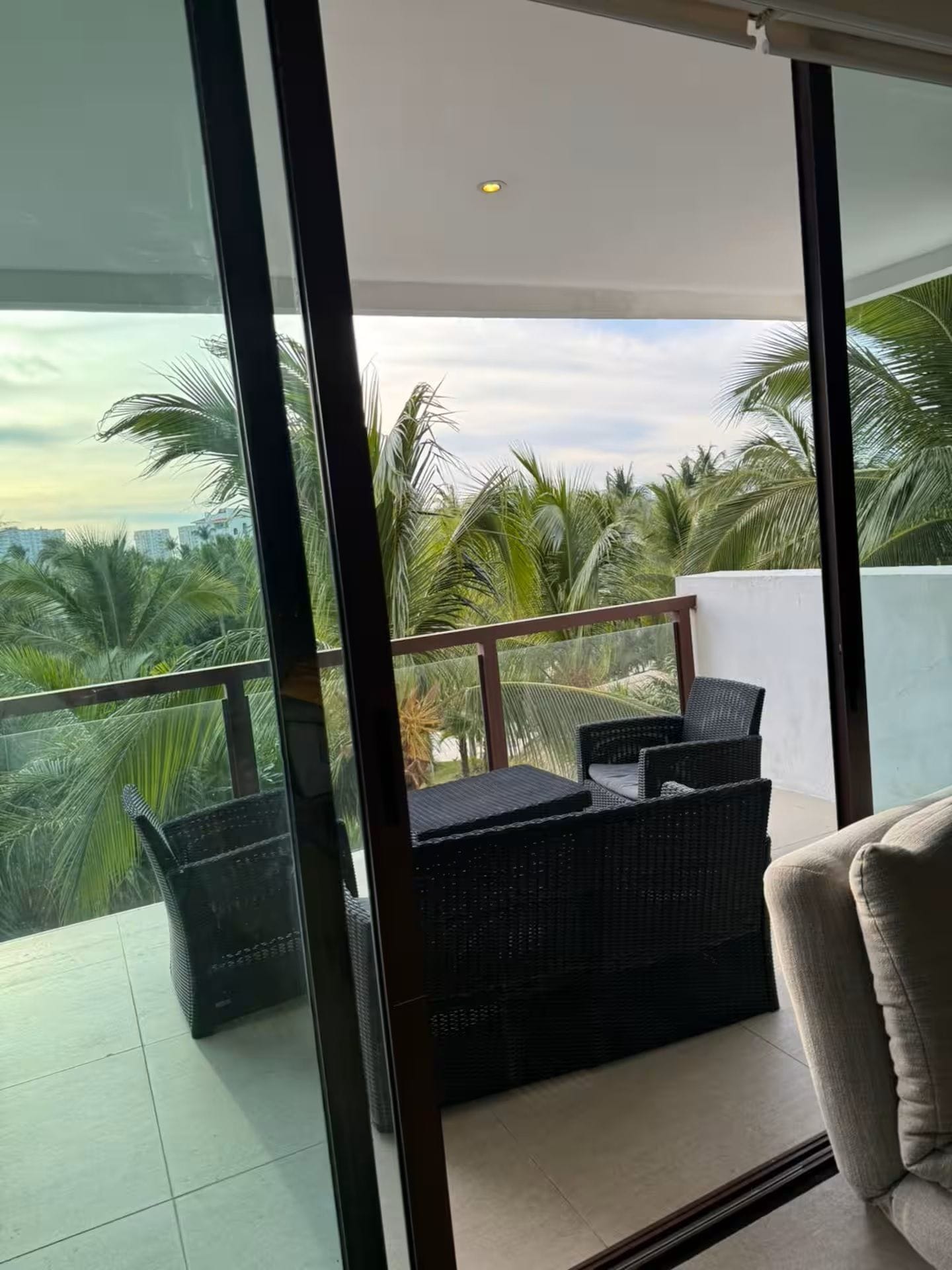 A balcony with a wicker sofa and a chair, overlooking a palm tree garden.
