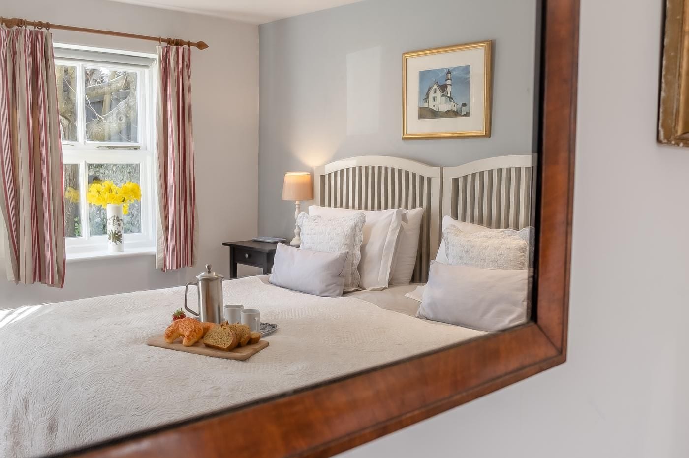 A neatly made bed with pillows and a tray of bread, a bedside table with a lamp, and a window with curtains.