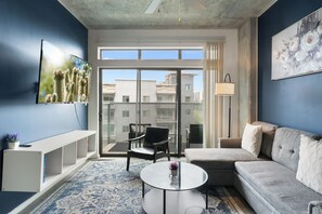 Ein modernes Wohnzimmer mit einer Couch, einem Couchtisch und einem Sessel. Ein großes Fenster bietet Blick auf Gebäude.
