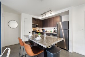 Eine moderne Küche mit einem Edelstahl-Kühlschrank, einer Mikrowelle und einer Arbeitsplatte aus Granit. Zwei braune Stühle und ein Mülleimer sind ebenfalls vorhanden.