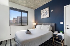 A hotel room with a large bed, a bedside table with a lamp, a small plant, and a view of buildings through the window. - Luxury 2BR | Pool + W/D | Near Arts District (Phoenix)