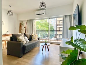 Ein modernes Wohnzimmer mit einer Couch, einem kleinen runden Tisch und einem großen Fenster mit Blick auf einen Balkon und Gebäude.