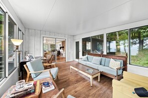 A spacious living room with a sofa, coffee table, and chairs, large windows offering a view of the outdoors. - Stunning Crystal Lake Views | Private Hot Tub | Sleeps 10 | Near Beulah (Beulah)