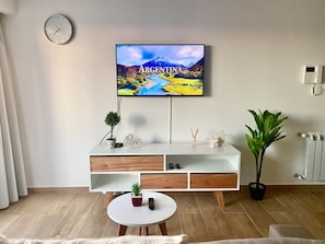 Ein modernes Wohnzimmer mit einem Flachbildfernseher, der ein Landschaftsbild von Argentinien zeigt, einem weißen Konsolentisch mit Schubladen, einem kleinen runden Tisch mit einer Pflanze und einem Topfpflanzen neben dem Tisch.
