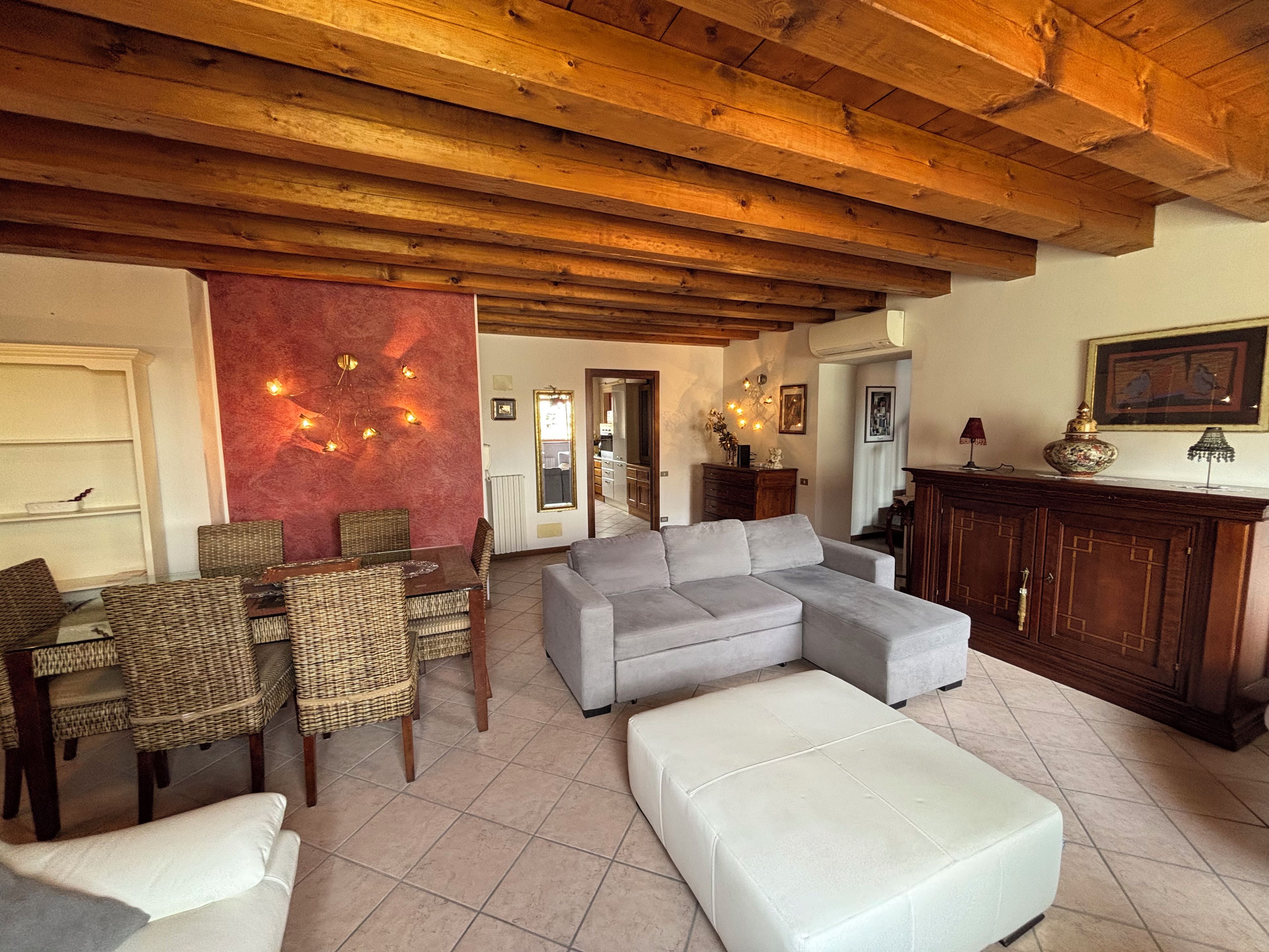 A living room with a sectional sofa, a dining table with wicker chairs, a fireplace, and a kitchen area visible through an open doorway.