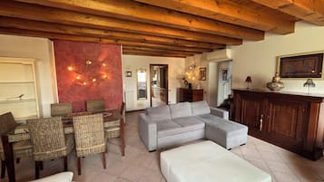 A living room with a sectional sofa, a dining table with wicker chairs, a fireplace, and a kitchen area visible through an open doorway.