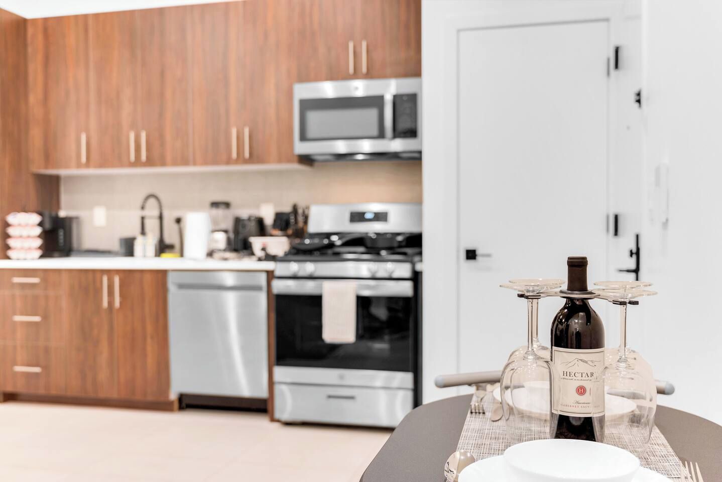 A modern kitchen with wooden cabinets, stainless steel appliances, and a dining area with a wine bottle and glasses.