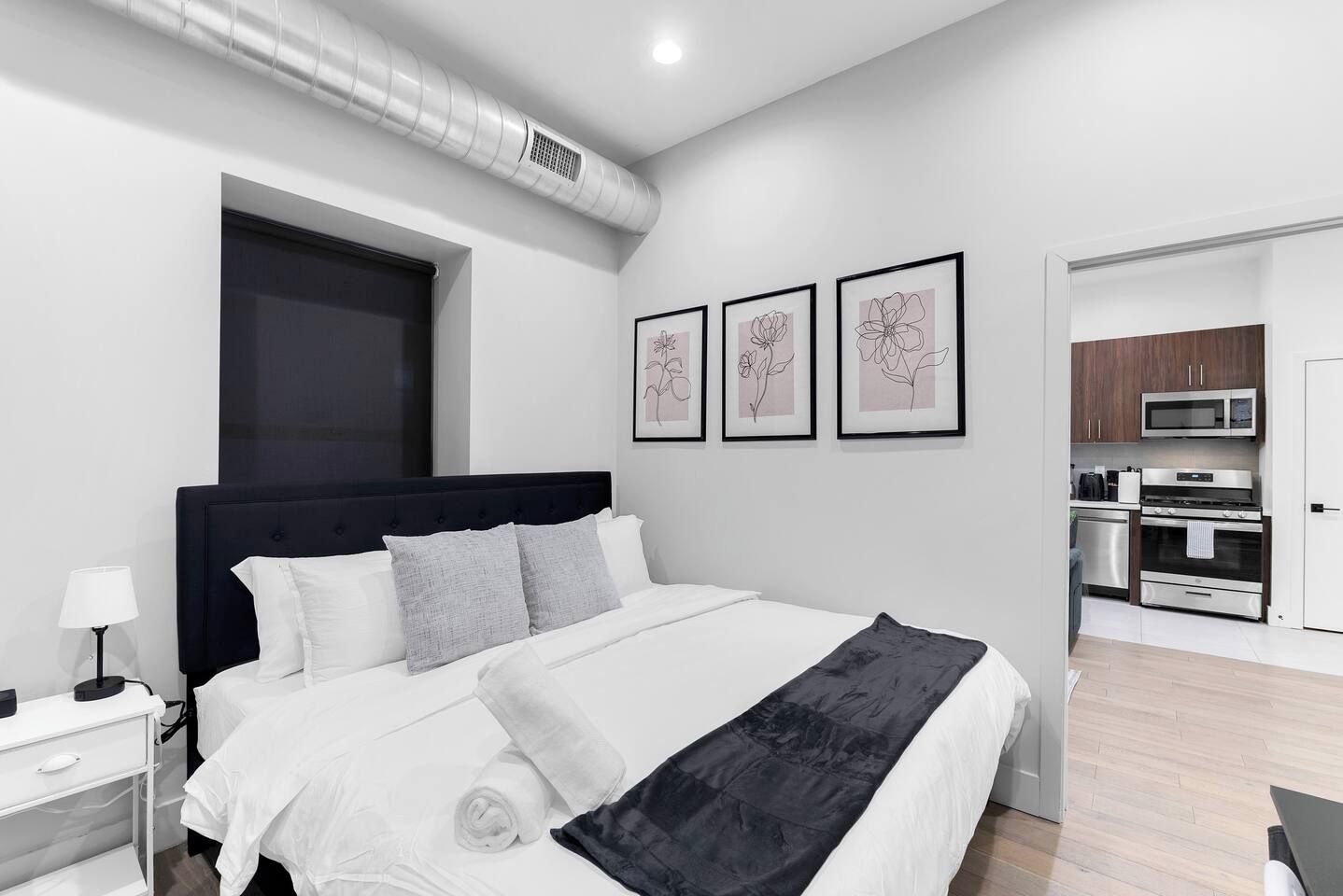 Une chambre d’hôtel moderne avec un grand lit, une table de chevet avec une lampe, une bouche d’aération fixée au mur et une vue sur la cuisine.