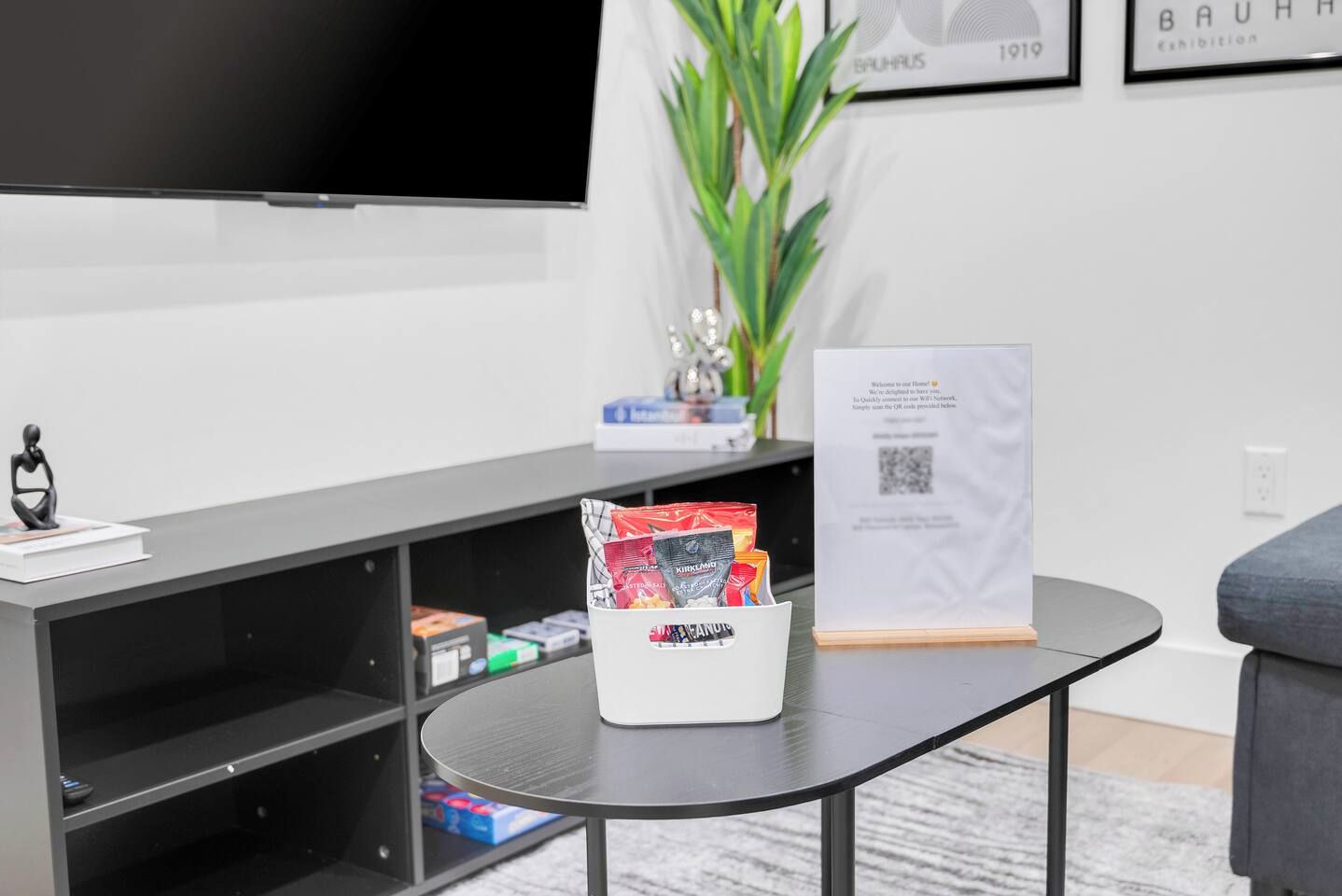 A modern living room with a flat-screen TV, a coffee table with snacks, a framed picture, and a plant.