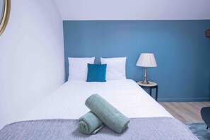 Ein Hotelzimmer mit einem Bett, einem Nachttisch, einer Lampe und einem zusammengerollten Handtuch auf dem Bett.