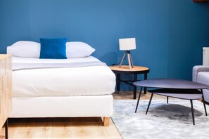 A bed with white bedding and a blue pillow, a small round table with a lamp, and a modern chair. - New Listing (Choisy-le-Roi)