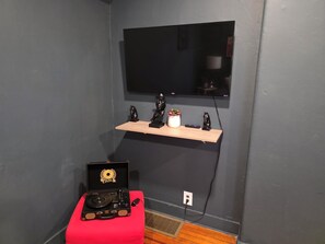 A room with a flat-screen TV mounted on the wall, a wooden shelf with decorative items, a red ottoman, and a vintage record player on a red cushion. - Enjoyable 4-bedroom house with AC in charming Meriden (Meriden)