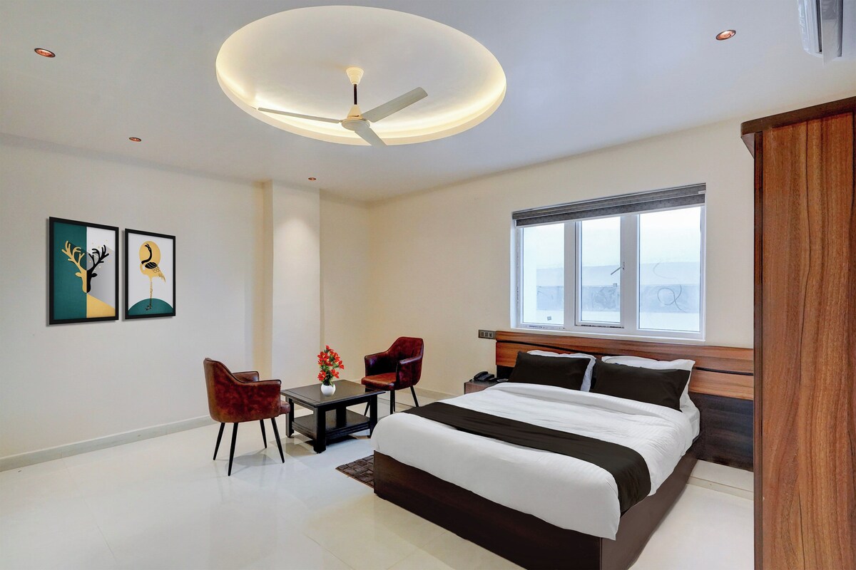 A modern hotel room with a large bed, a desk, two chairs, a ceiling fan, and a window with a view.