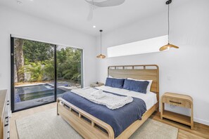 A modern bedroom with a large bed, a small bedside table, a nightstand, and a view of a swimming pool through a sliding glass door. - Sarasota Coastal Retreat (Sarasota)