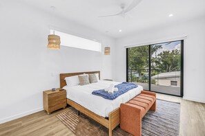 Een moderne slaapkamer met een groot bed, een houten hoofdbord, nachtkastjes, een plafondventilator en uitzicht op de natuur door een groot raam.
