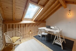 Ein gemütliches Dachzimmer mit Holzbalkendecke, einem Oberlicht, einem weißen Schreibtisch mit Laptop, einem Korbstuhl und einem kleinen Tisch.