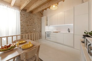 A modern kitchen with white cabinets, a stone wall, and a dining area with a table set for two. - Casinha do Lagar - Rustic Stay (Nazaré)
