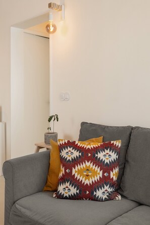 Ein graues Sofa mit gemusterten Kissen, ein kleiner Tisch mit einer Pflanze und eine wandmontierte Leuchte.