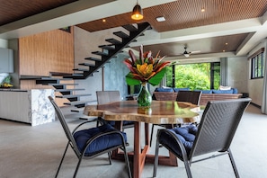 Ein modernes Interieur mit einem runden Holztisch, dunkel gepolsterten Stühlen, einer Treppe mit Holzhandläufen und einem großen Fenster mit Blick auf Grünflächen.