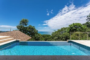 Piscina azul límpida com degraus que descem até a água, cercada por vegetação exuberante e com vista para um corpo d'água.