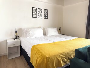 Ein ordentlich bezogenes Bett mit weißer Bettwäsche und einer gelben Tagesdecke, eine Nachttischlampe und ein Nachttisch mit einer Schublade.