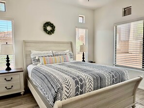 A bedroom with a bed, nightstands, a lamp, a wall-mounted wreath, and windows with blinds. - Vistoso Resort Condo #107A (Oro Valley)