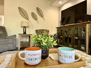A cozy living room with a sofa, a coffee table with two mugs, a potted plant, a lamp, and a TV on a cabinet. - Vistoso Resort Condo #107A (Oro Valley)