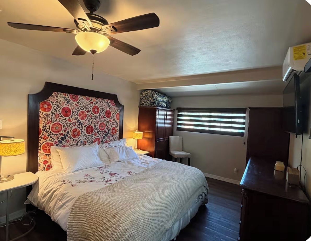 A bedroom with a bed, a ceiling fan, a window with blinds, a nightstand, and a lamp.