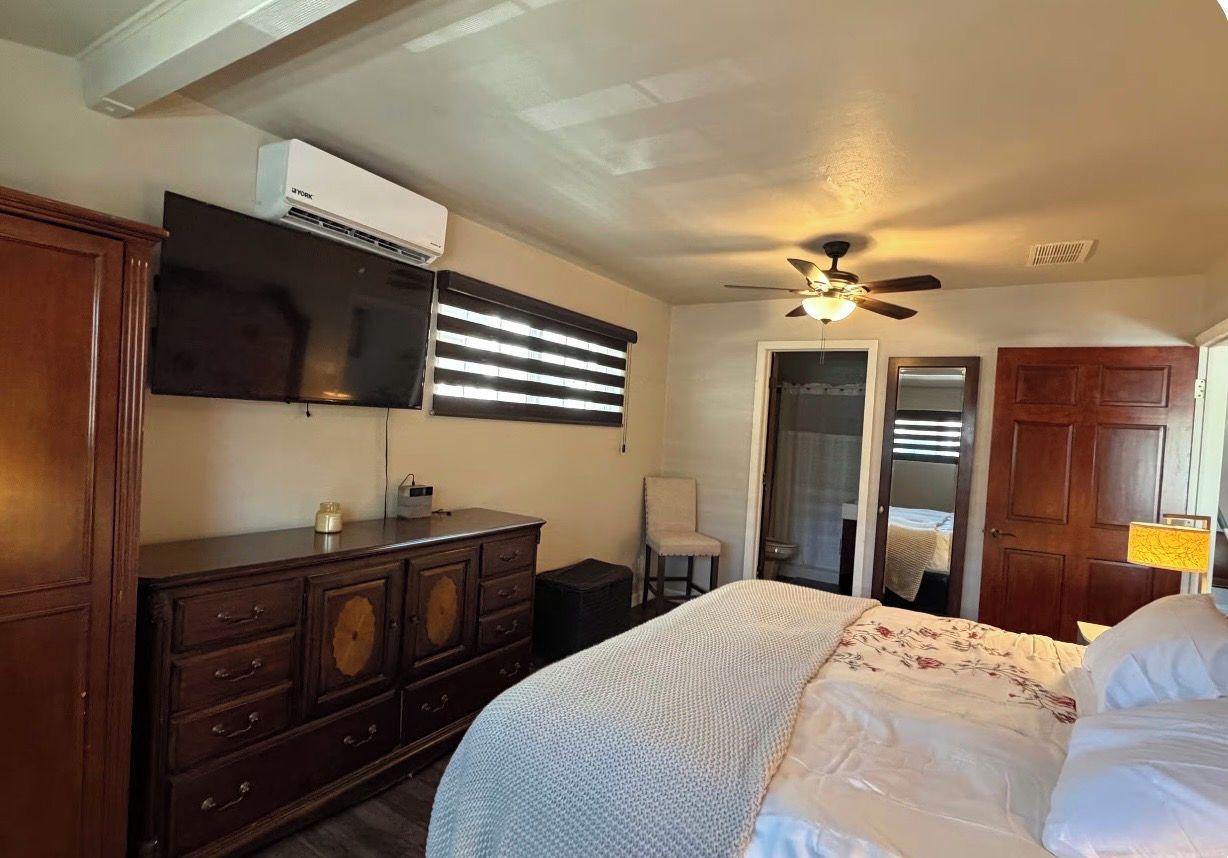 A bedroom with a bed, dresser, TV, ceiling fan, and a door leading to another room.