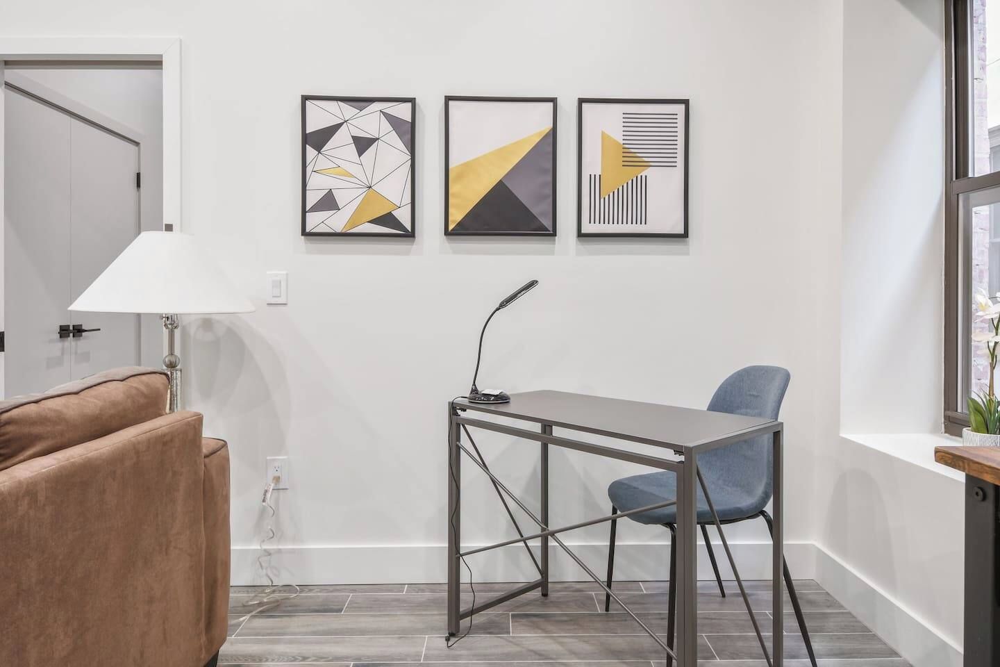 A modern room with a desk, chair, and a lamp. There are three framed abstract artworks on the wall.