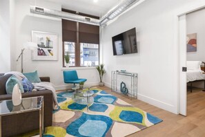 A modern living room with a sofa, a blue chair, a glass coffee table, and a colorful rug. There is a television mounted on the wall and a window with blinds. - 1BR 20 Min Manhattan 2 min prud center. Concerts! (Newark)