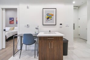 Een moderne, minimalistisch ingerichte kamer met een bed, een bureau en een stoel. Op de muur hangt een lijst met een abstract schilderij, op het bureau staat een kleine potplant en er is een prullenbak naast het bureau.