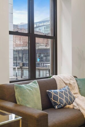 A room with a large window, a brown sofa with patterned cushions, and a view of a building with 'Prudential' signage. - 1BR 20 Min Manhattan 2 min prud center. Concerts! (Newark)