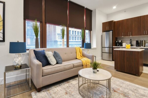 A modern living room with a sofa, a coffee table, and a kitchen area with wooden cabinets and appliances. - Charming 1BR King Bed - Just 20 Minutes from NYC! (Newark)