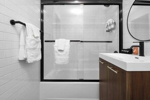 A modern bathroom with a glass-enclosed shower, white tiled walls, and a wooden vanity with a mirror. - Charming 1BR King Bed - Just 20 Minutes from NYC! (Newark)