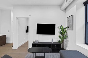 A modern living room with a flat-screen TV mounted on the wall, a black entertainment unit, a grey sofa, and a dining table with chairs. - 1BR King 20 min from NYC! 2 minutes to prudential (Newark)
