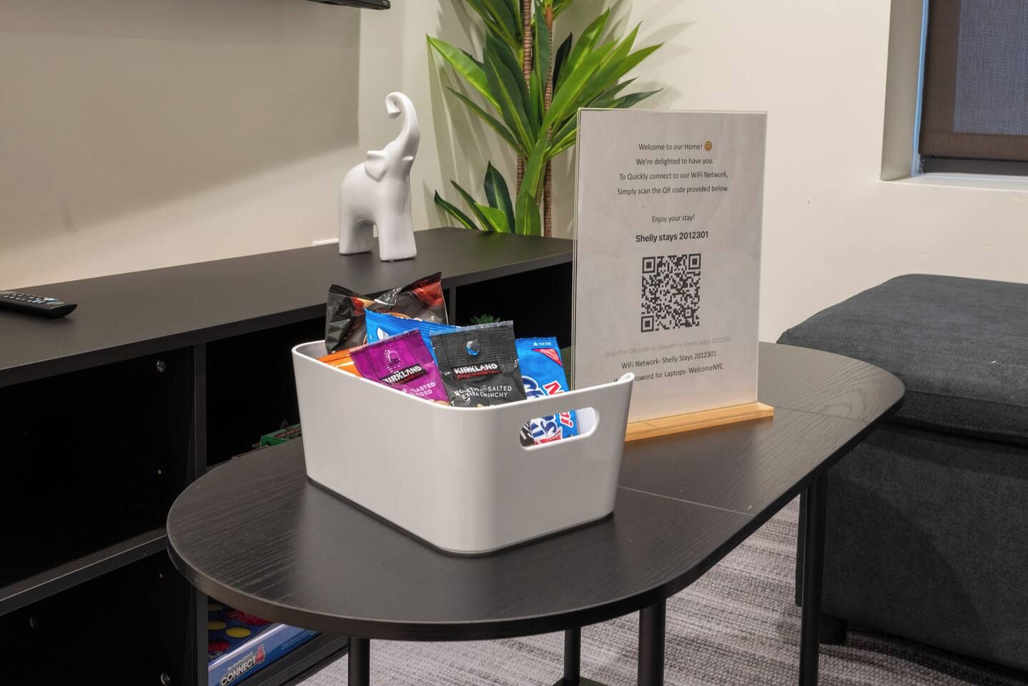 Ein Hotel-Rezeptionsbereich mit einer weißen Mülltonne, gefüllt mit Snacks, einem QR-Code-Schild für WLAN-Zugang, einer Fernbedienung und einer kleinen weißen Elefantfigur auf einem schwarzen Tisch.