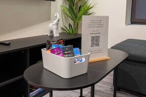 Zona de recepción de un hotel con un cubo blanco lleno de paquetes de comida, un cartel con código QR para acceso a WiFi, un control remoto y una pequeña figurita de elefante blanco sobre una mesa negra.