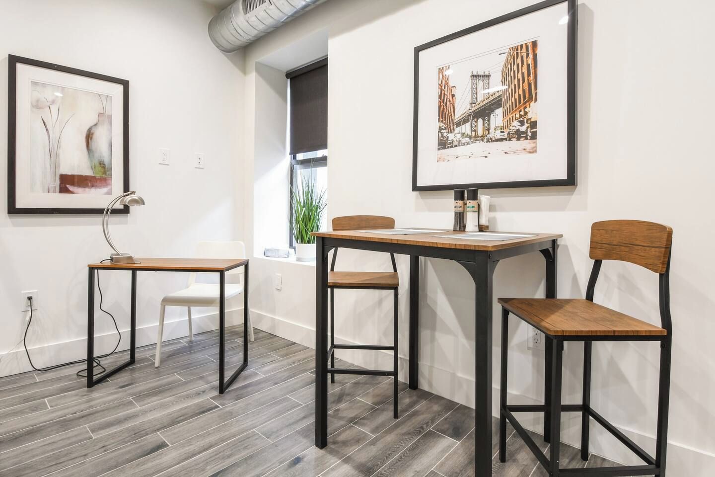 A modern room with a wooden table and chairs, a desk with a lamp, and two framed pictures on the wall.