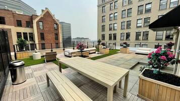 An outdoor patio with a wooden table and benches, surrounded by planters with flowers, and a trash bin.