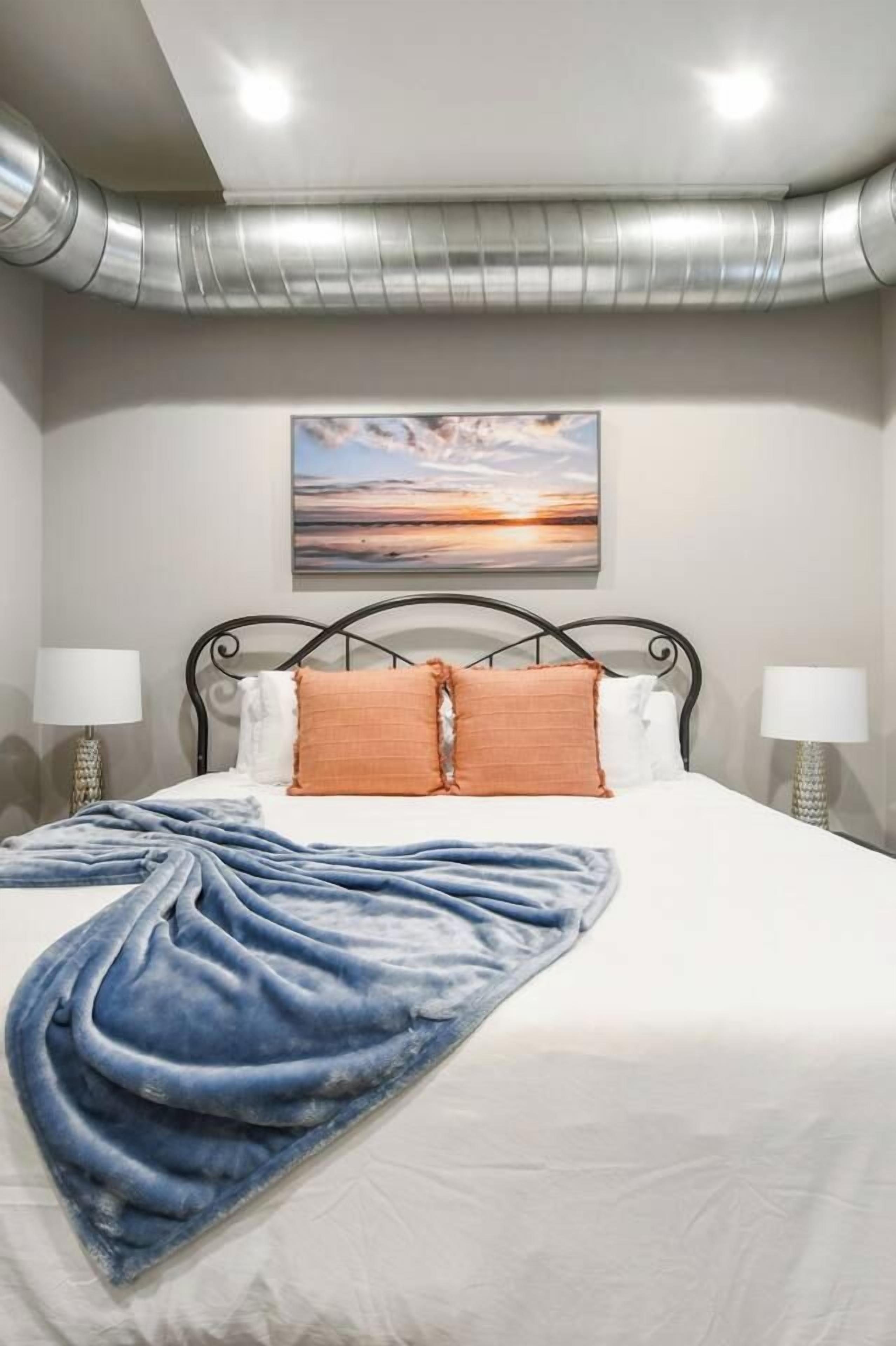 A hotel room with a bed, two bedside lamps, a painting on the wall, and a ceiling vent.