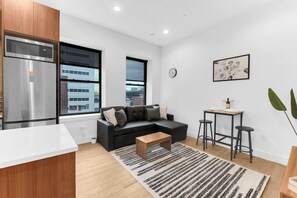 A modern kitchen with a built-in microwave and refrigerator, a black leather sofa with a coffee table, a small bar area with stools, and large windows offering a city view. - 1BR Oasis, 20 Min NYC, 4 Min to Prudential Concert (Newark)