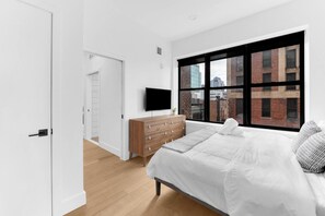 A modern bedroom with a large bed, a wooden dresser, a television, and a window with a city view. - 1BR Oasis, 20 Min NYC, 4 Min to Prudential Concert (Newark)
