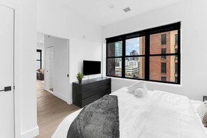 A modern bedroom with a large window, a flat-screen TV, a black dresser, and a bed with white linens. - Monthly friendly Lux 1BR 20 Min NYC 4 min to Prud (Newark)