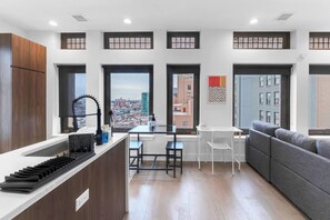 A modern kitchen with a central island, a dining area with a white table and chairs, and a grey sofa. - 20 Min to NYC Modern Lux Escape– 1BR, Gym rooftop (Newark)