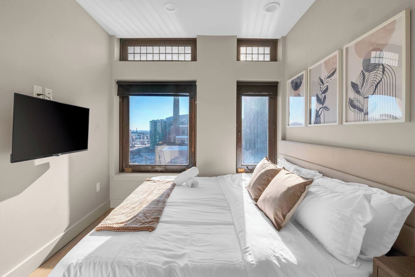 A bedroom with a large bed, two windows, a flat-screen TV, and artwork on the wall.