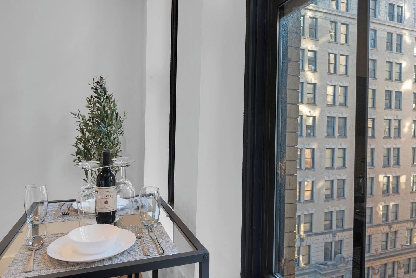 A dining table set with a bottle of wine, glasses, and a small plant, with a view of a building through a window.