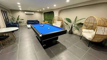 A pool table in a room with a tiled floor, a sofa, and two wicker chairs.