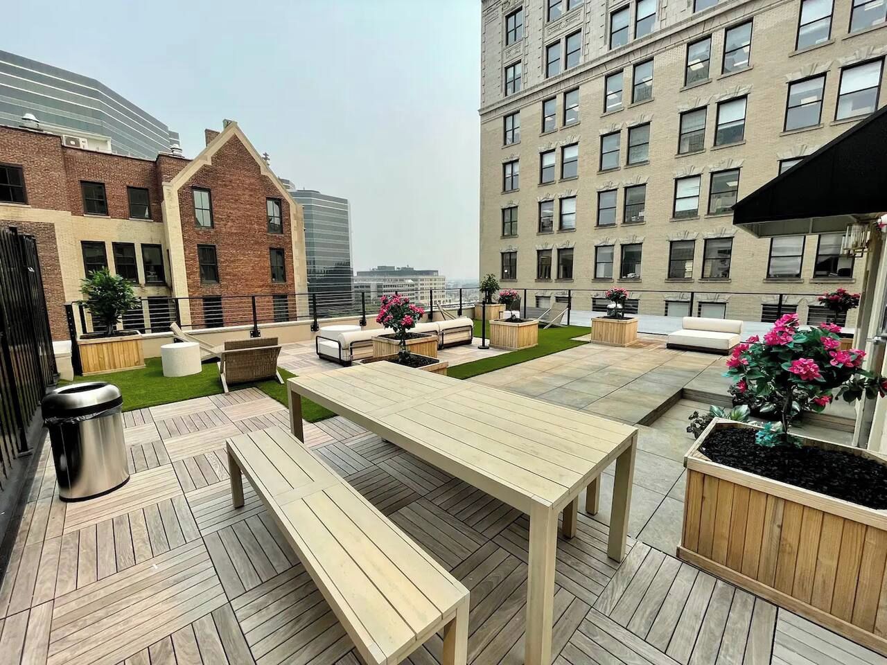 An outdoor patio with a wooden table and benches, surrounded by planters with flowers, and a trash bin.